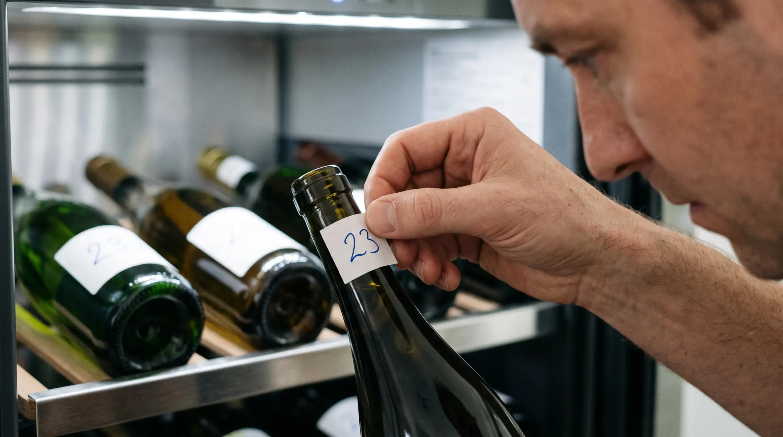 Main d'adulte apposant une étiquette autocollante numérotée sur le goulot d'une bouteille de vin, avec d'autres bouteilles déjà étiquetées visibles sur une clayette de cave en arrière-plan